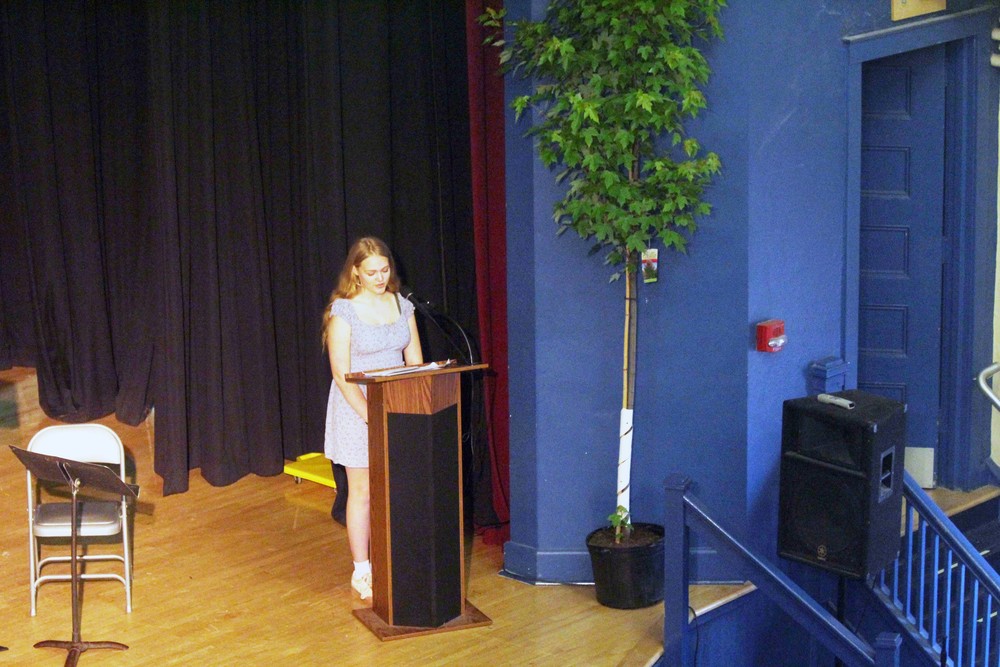 girl reading from lectern while standing next to tree in pot on stage