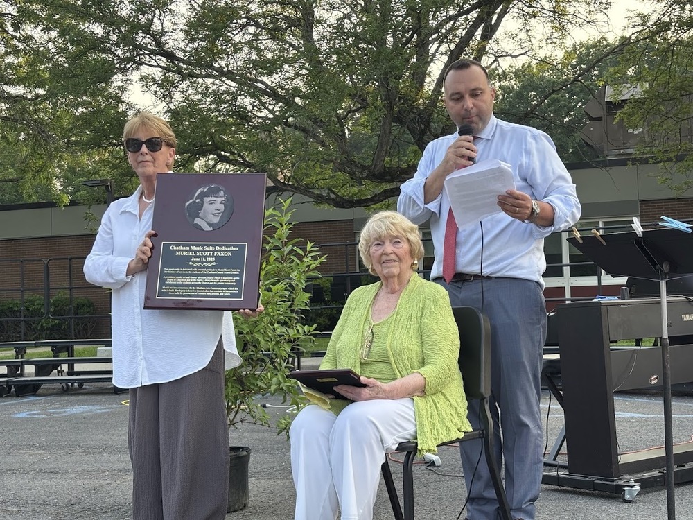 woman holding plaque next Ms. Faxon seated in chair with man standing behind her reading a speech